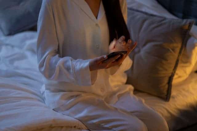 woman in pyjamas with smartphone in hand sits on bed