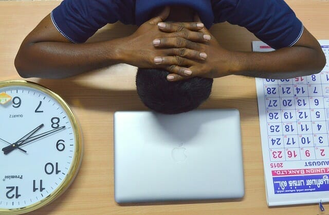 Overtired man keeps his head on a desk, next to a laptop, calendar, clock